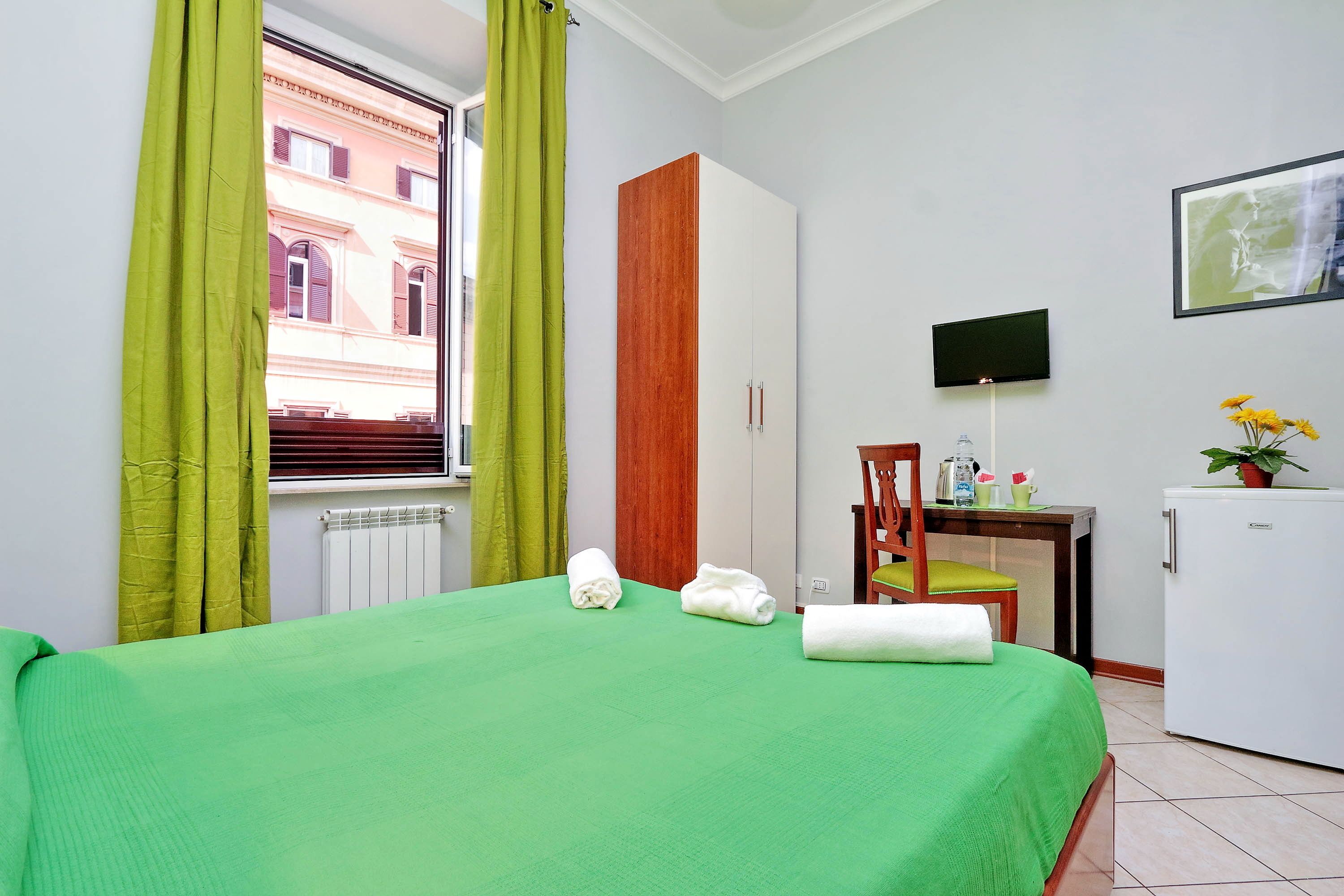 Habitación de hotel con colcha verde, armario de madera, escritorio con silla, refrigerador pequeño y televisor en la pared.