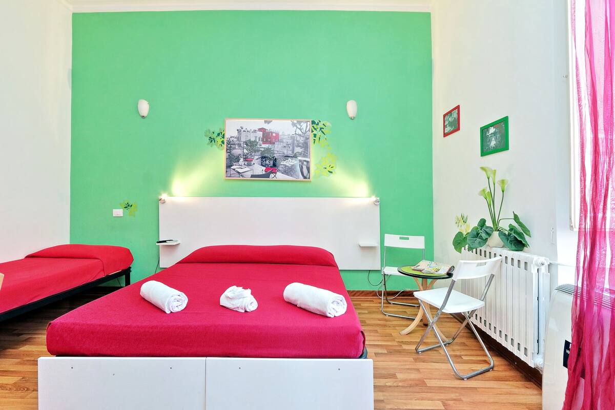 Camera d'albergo con parete verde, letto rosso e termosifone bianco. Ci sono due asciugamani bianchi sul letto e un piccolo tavolo con una sedia.