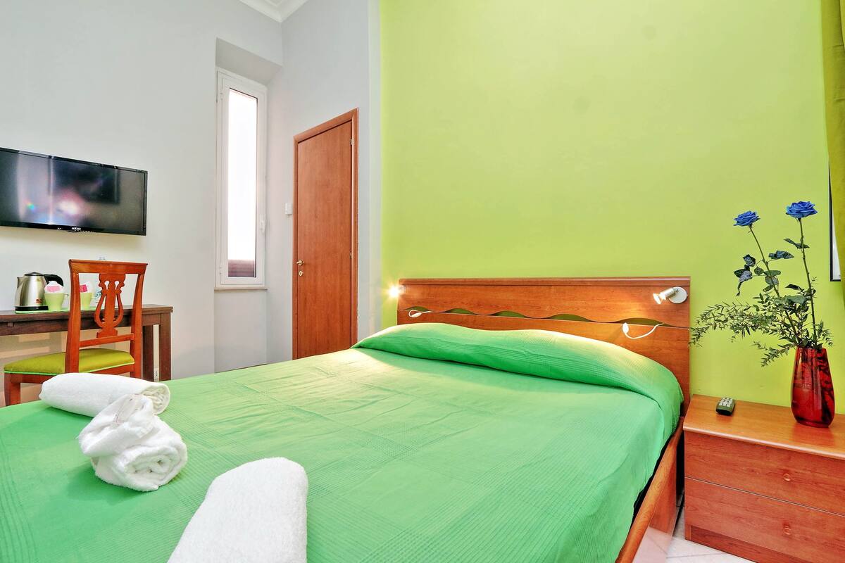 Un letto rifatto con lenzuola verdi, una testiera in legno, un comodino di legno con un vaso di fiori blu e un televisore appeso al muro.