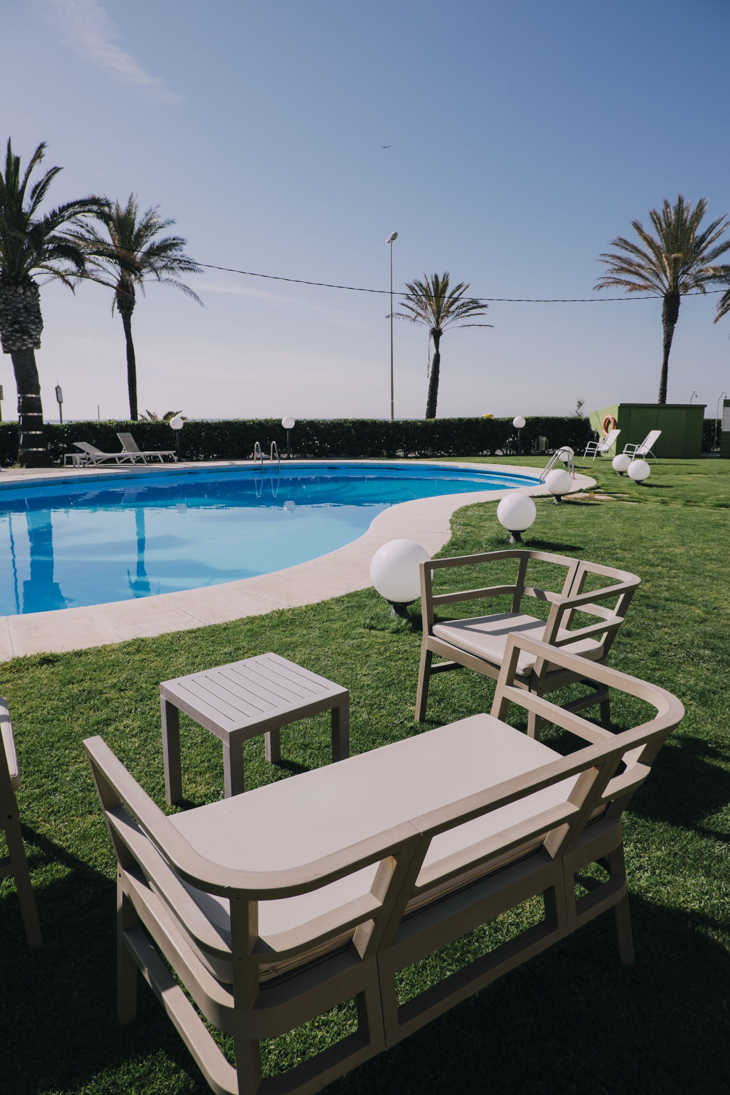 Un área de piscina con sillones de descanso y una mesa, rodeada de palmeras.