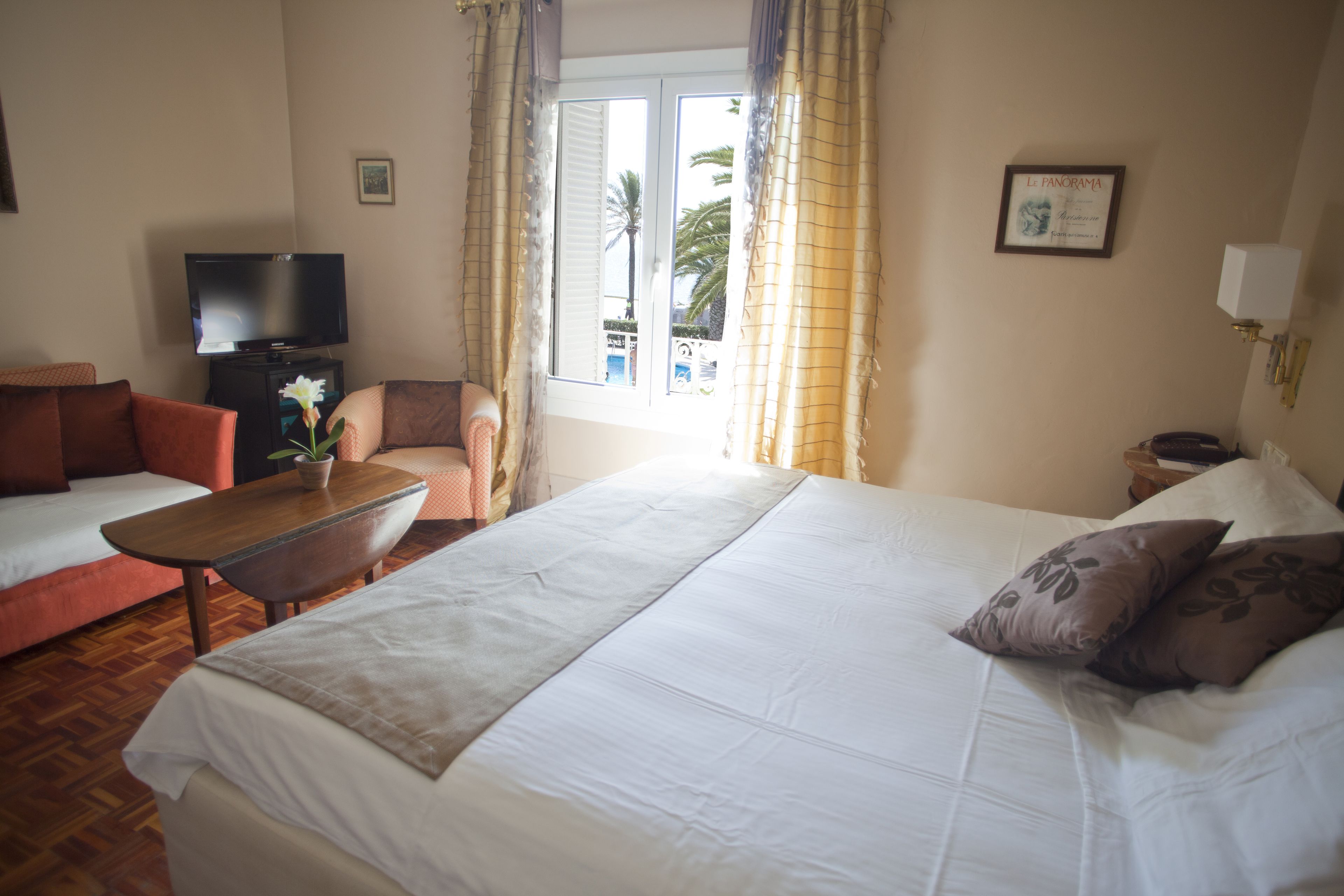 A hotel room with a bed, a sofa, a TV, a small table with a flower, and a window with curtains.