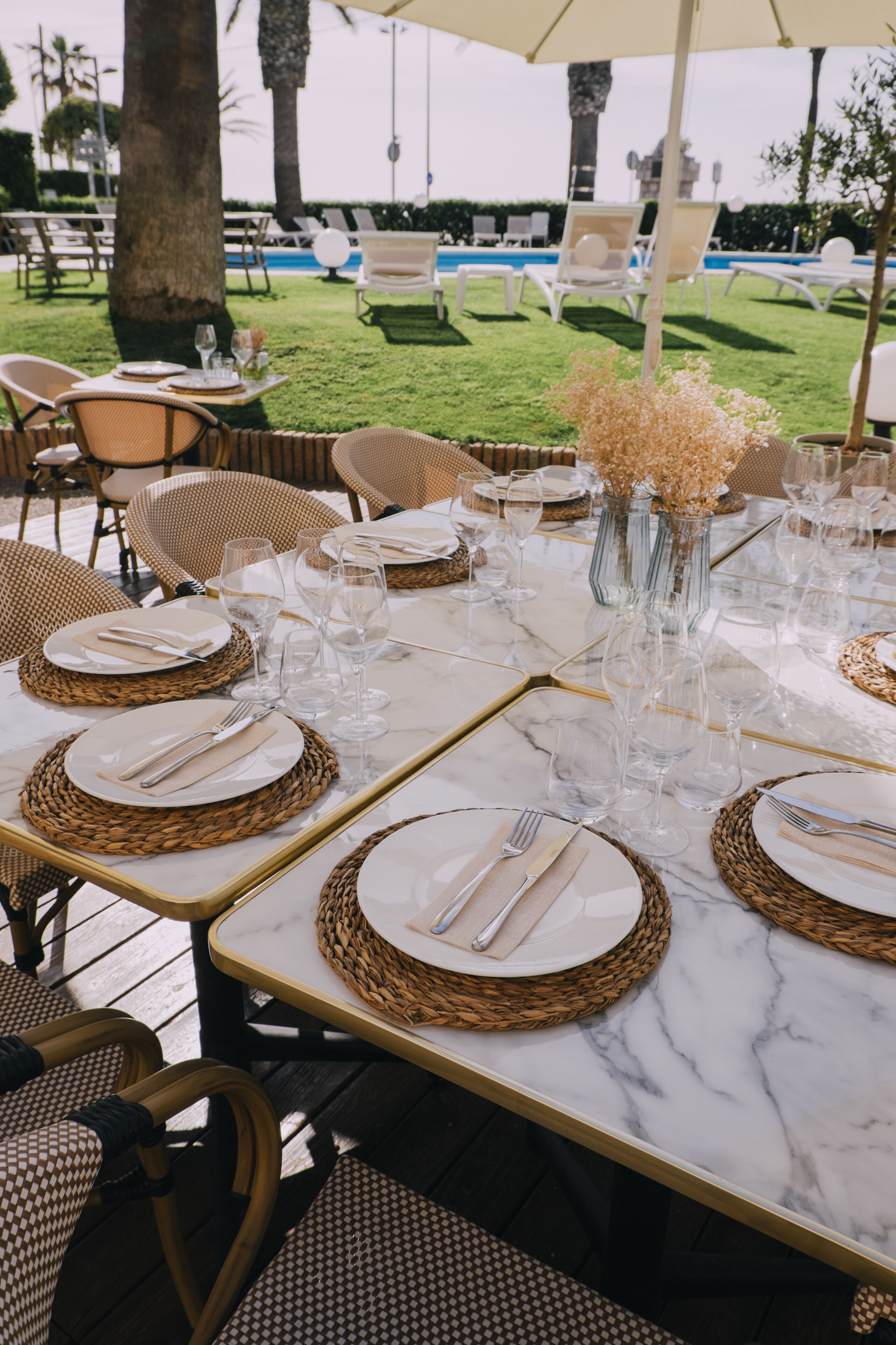 Una mesa preparada para una comida al aire libre, con individuales de mimbre, copas de cristal transparente y un jarrón con flores secas.