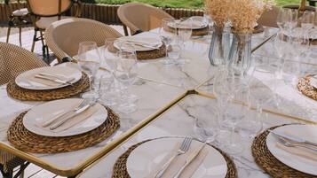 Una mesa preparada para una comida al aire libre, con individuales de mimbre, copas de cristal transparente y un jarrón con flores secas.