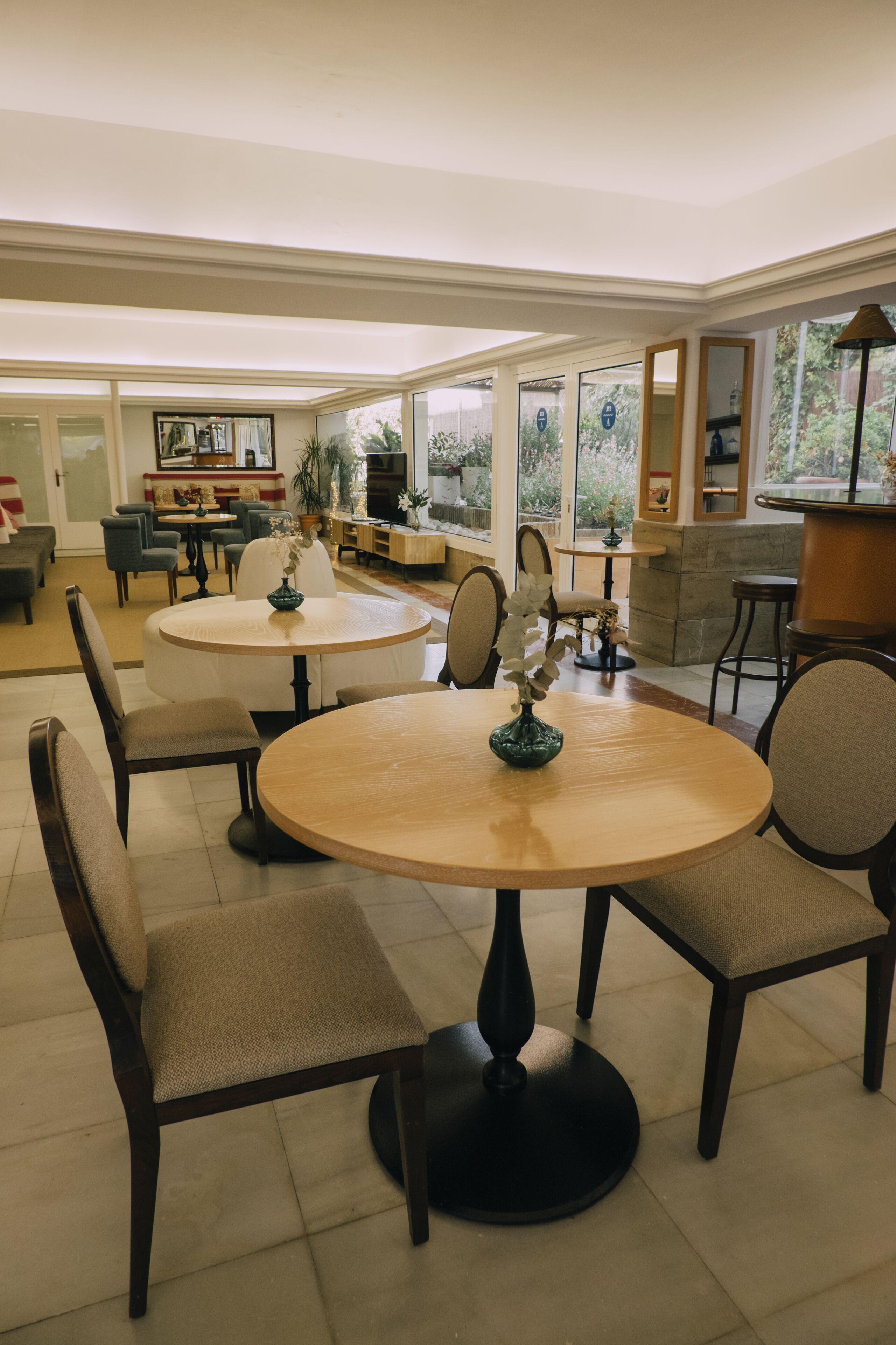 Un comedor amplio con mesas y sillas redondas, una barra de café y vistas al jardín a través de amplios ventanales.