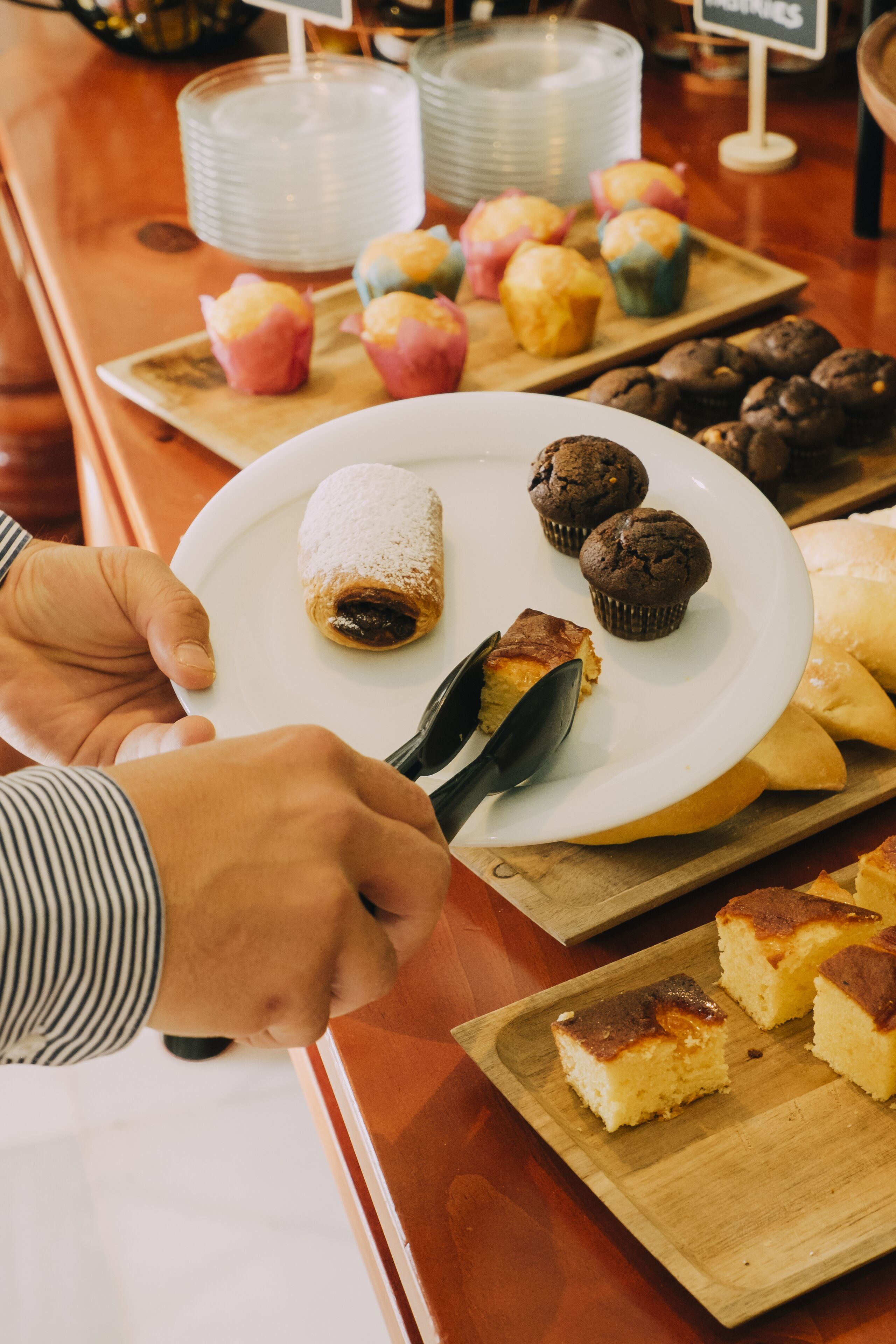 A person holding a plate with pastries and a spoon, with more pastries and plates in the background.