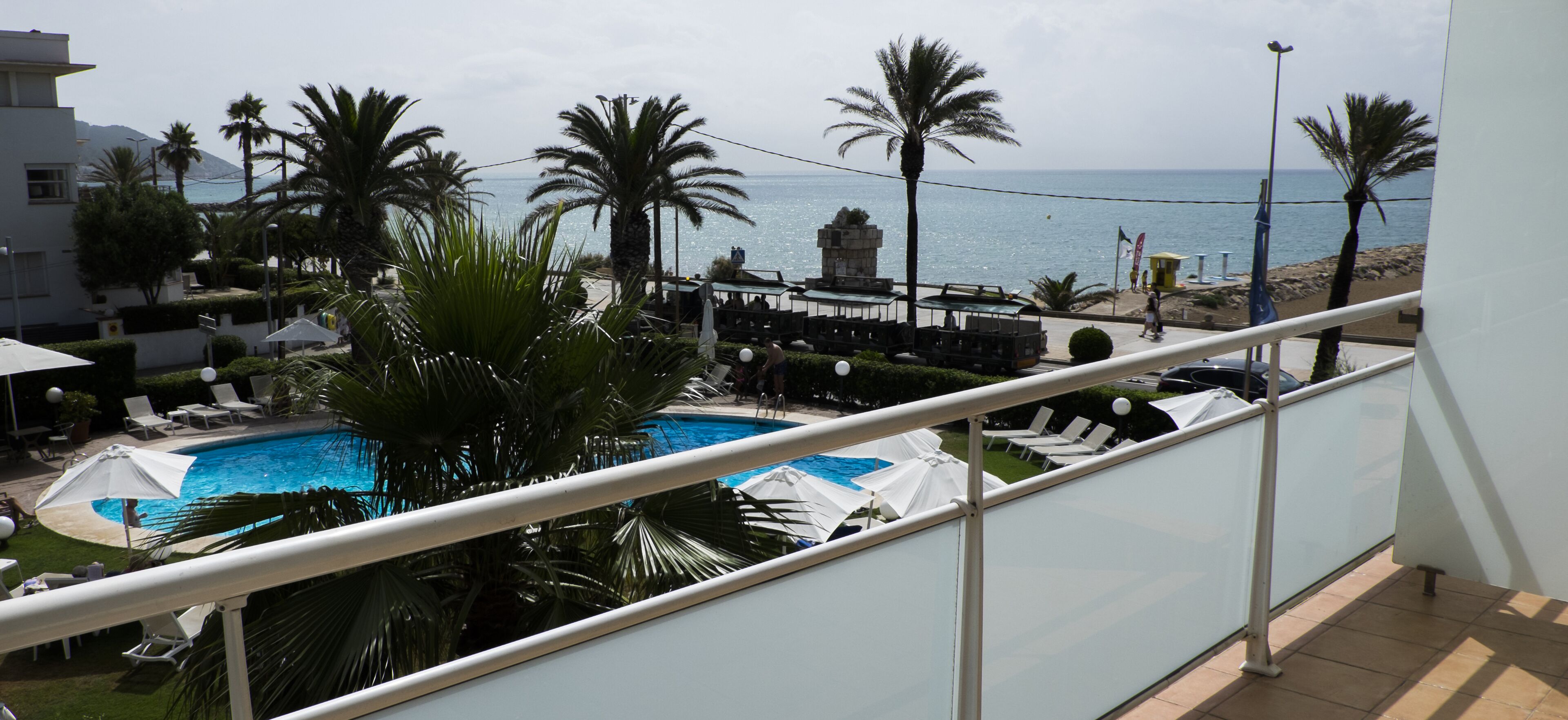 A pool area with lounge chairs and umbrellas, palm trees, and a view of the sea.