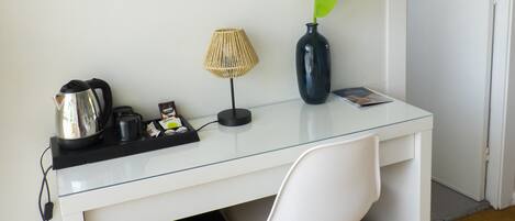 Una habitación compacta y moderna con un escritorio blanco, una silla, una obra de arte con una hoja enmarcada, un adorno verde, una lámpara, un mueble negro pequeño y un cubo de basura.