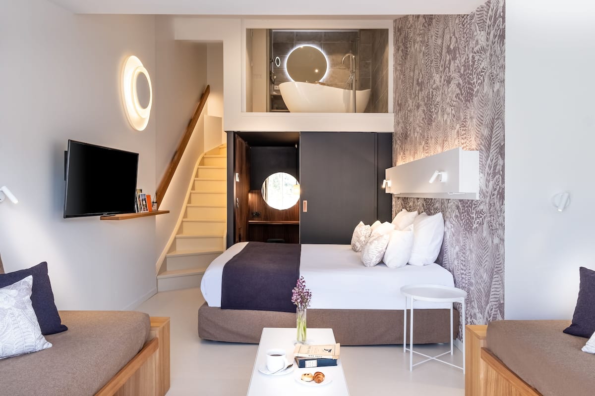 Habitación de hotel moderna con una cama grande, televisor de pantalla plana, una pequeña mesa con un jarrón y libros, y una escalera que lleva a un nivel superior con un baño.