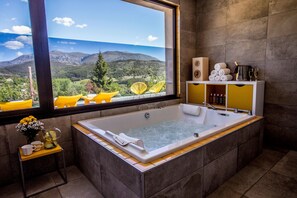 Une salle de bain moderne dotée d’une grande baignoire, d’une fenêtre offrant une vue panoramique sur les montagnes, ainsi que d’une petite table sur laquelle sont posées des serviettes et des articles de toilette.