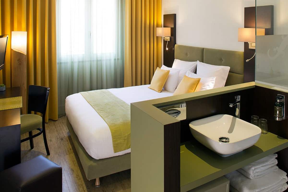 Habitación de hotel con cama, mesita de noche, lámpara, silla y baño con lavamanos y ducha.