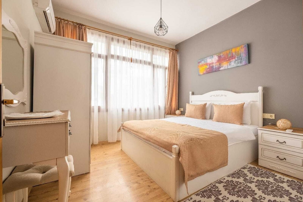 Ein Schlafzimmer mit Bett, Nachttisch, Kommode, Sessel, Fenster mit Vorhängen und einem Bild an der Wand.