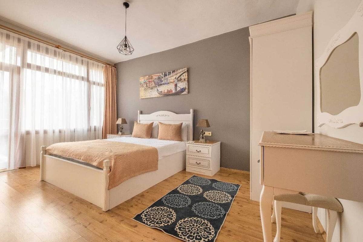 Ein Schlafzimmer mit einem Bett, Nachttischen, einer Kommode, einem Spiegel, einem gemusterten Teppich und einem Bild an der Wand.