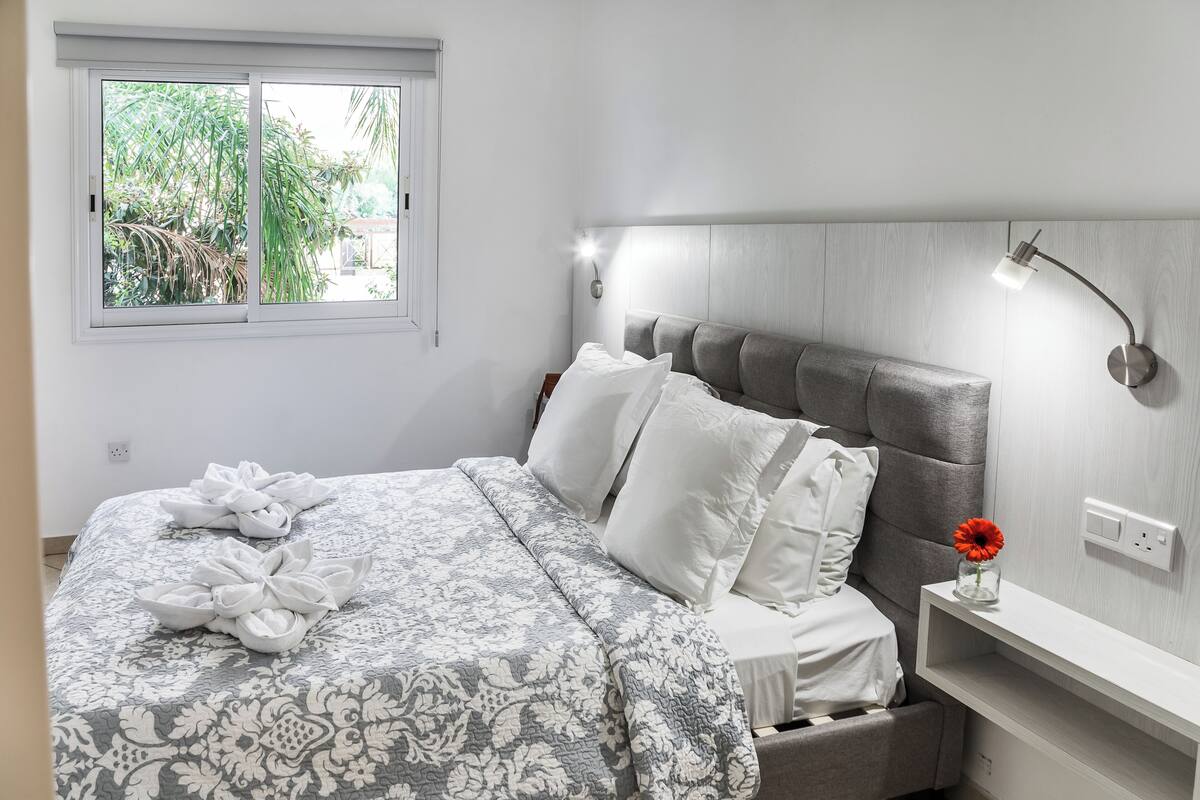 Uma cama bem arrumada com colcha estampada, uma mesa de cabeceira com abajur e uma janela com vista para área verde.