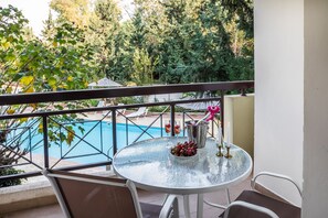 A balcony with a glass table, chairs, a metal railing, and a view of a swimming pool and trees.