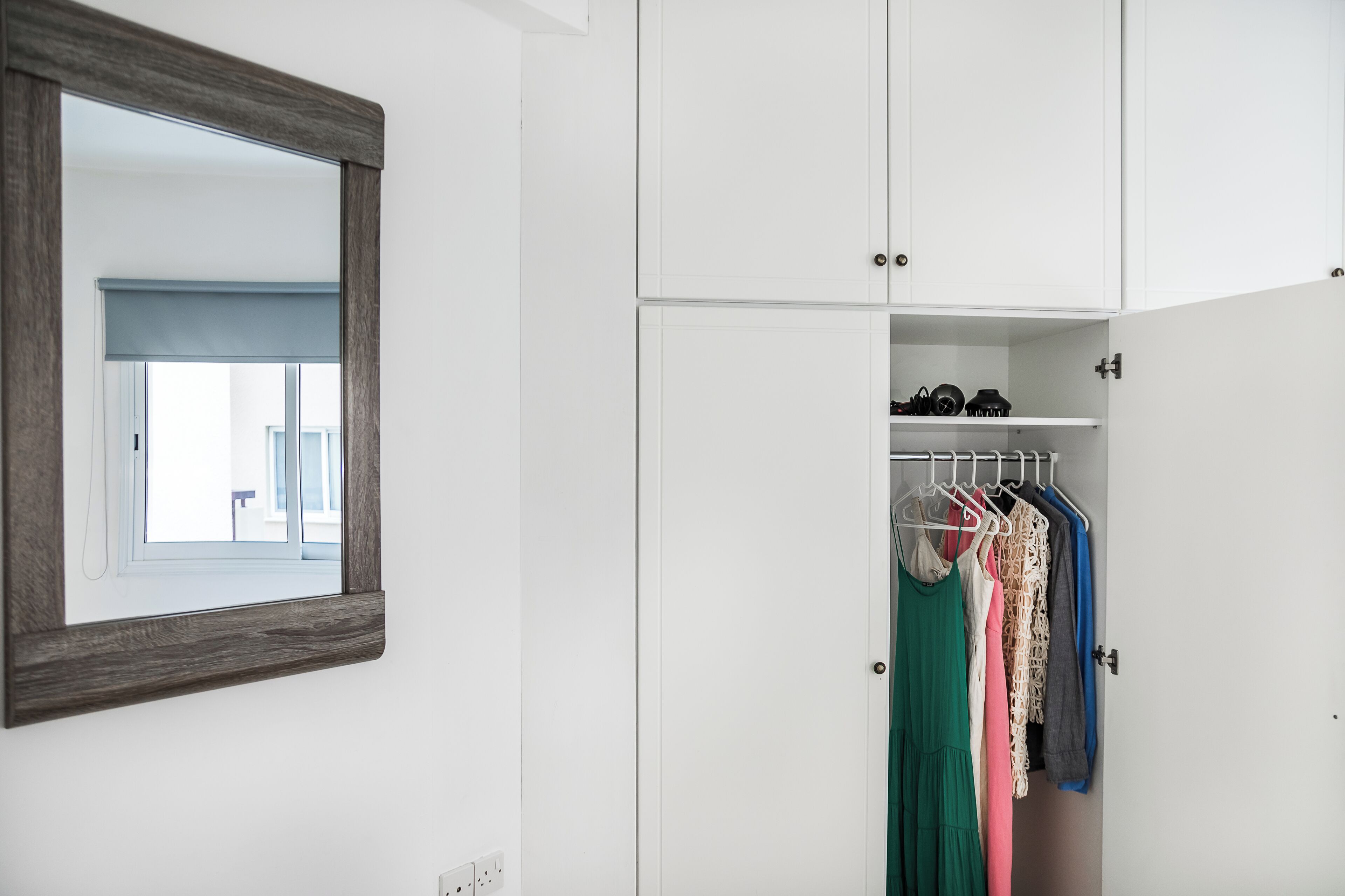 A white wardrobe with a mirror above it, a hanging rack with clothes, and a window with blinds.