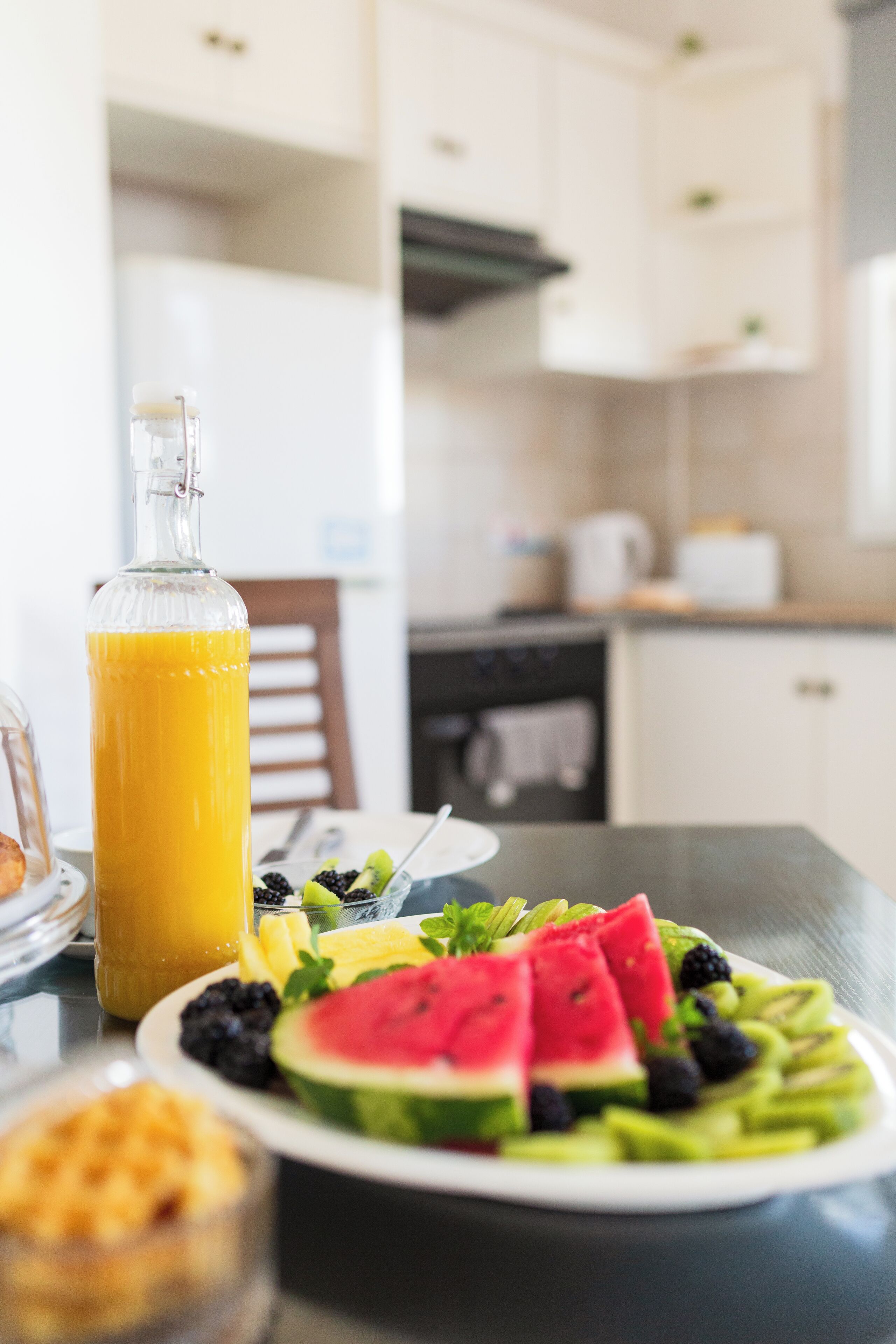 A breakfast table with a glass bottle of orange juice, a plate of fresh fruit, and a small bowl of cereal.