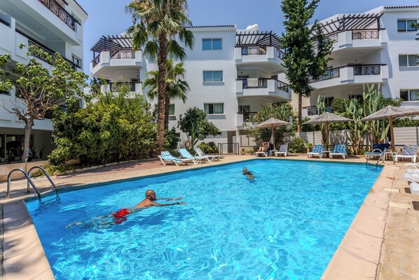 A swimming pool with lounge chairs and palm trees, surrounded by a multi-story building with balconies.