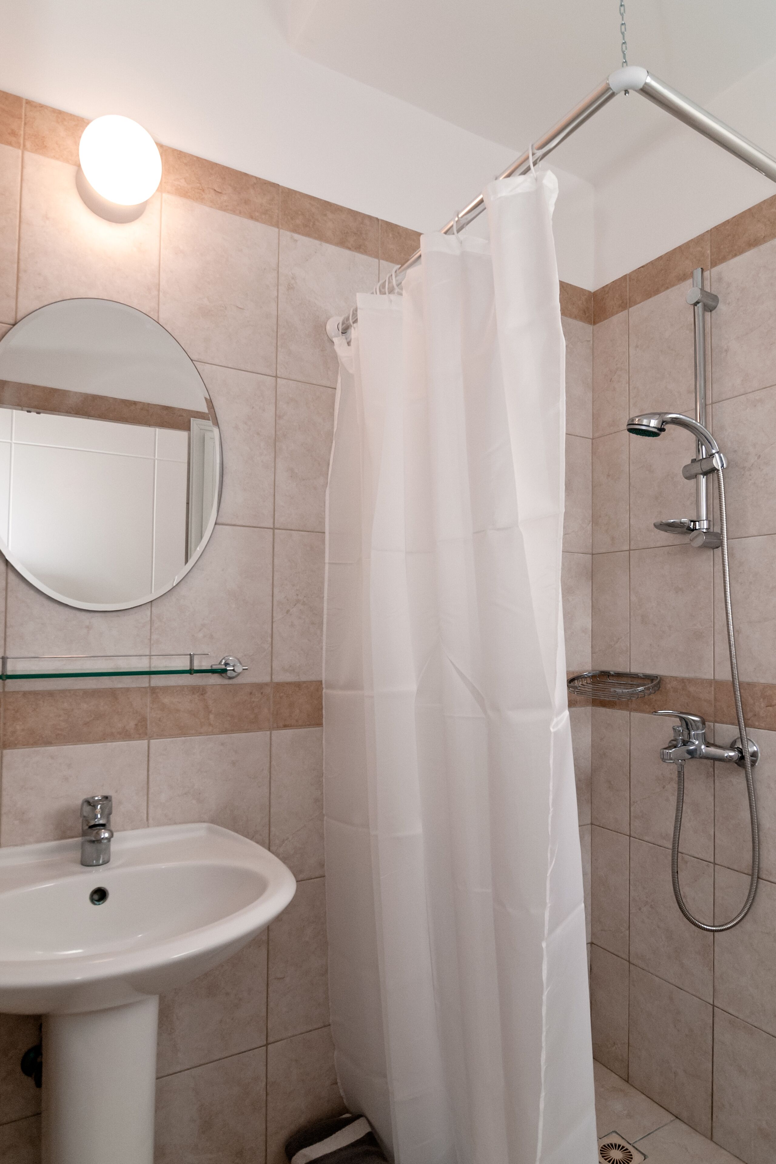 A bathroom with a white sink, a round mirror, a shower with a handheld showerhead, and a glass shelf.