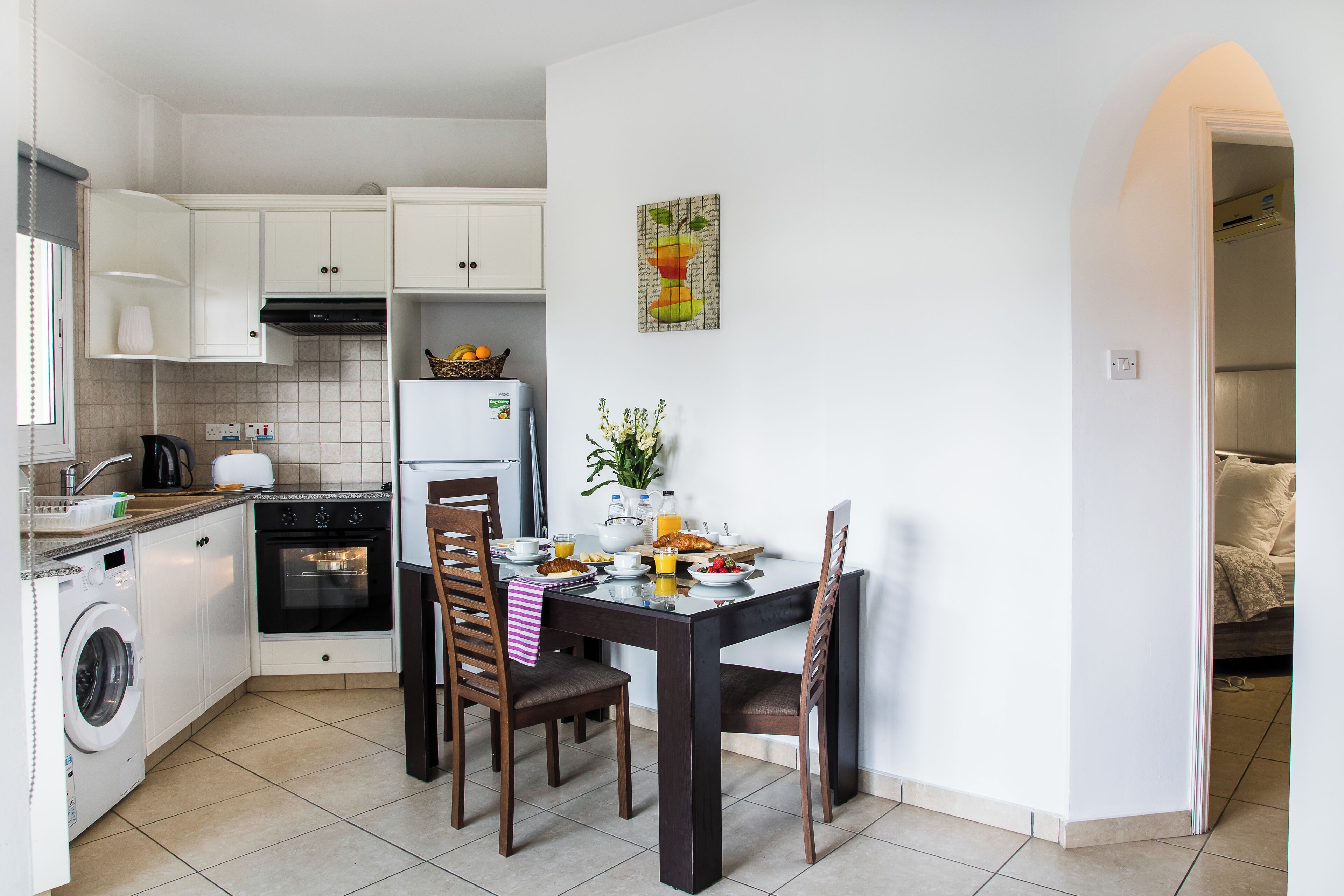 Una cocina compacta con armarios blancos, lavadora, horno y una mesa de comedor con sillas.