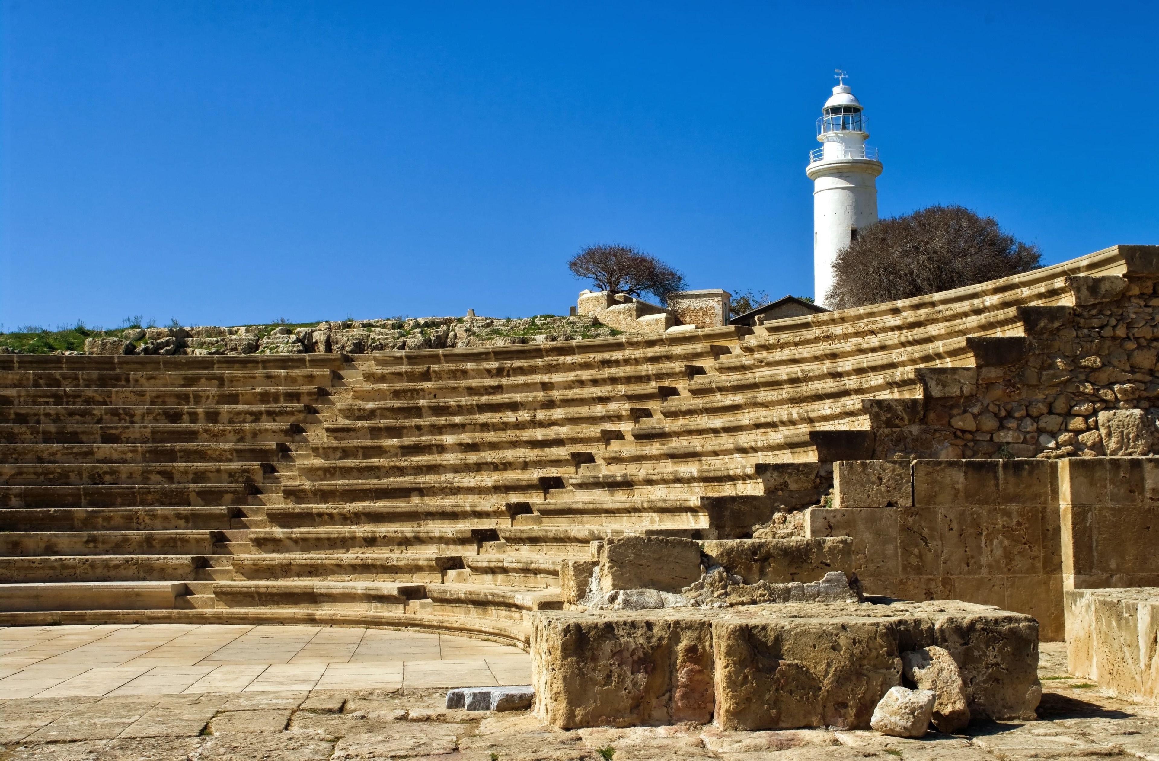 An ancient amphitheater with stone steps and a lighthouse in the background.