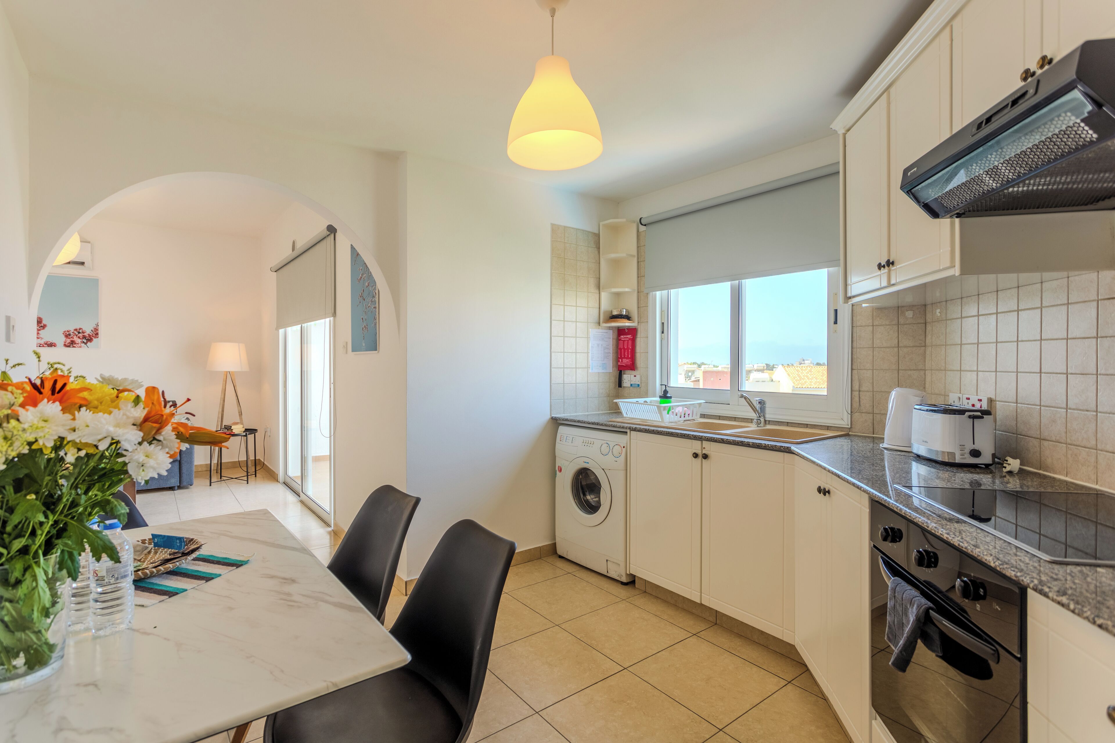 A modern kitchen with a washing machine, a dining area with a table and chairs, and a view of the city through the window.