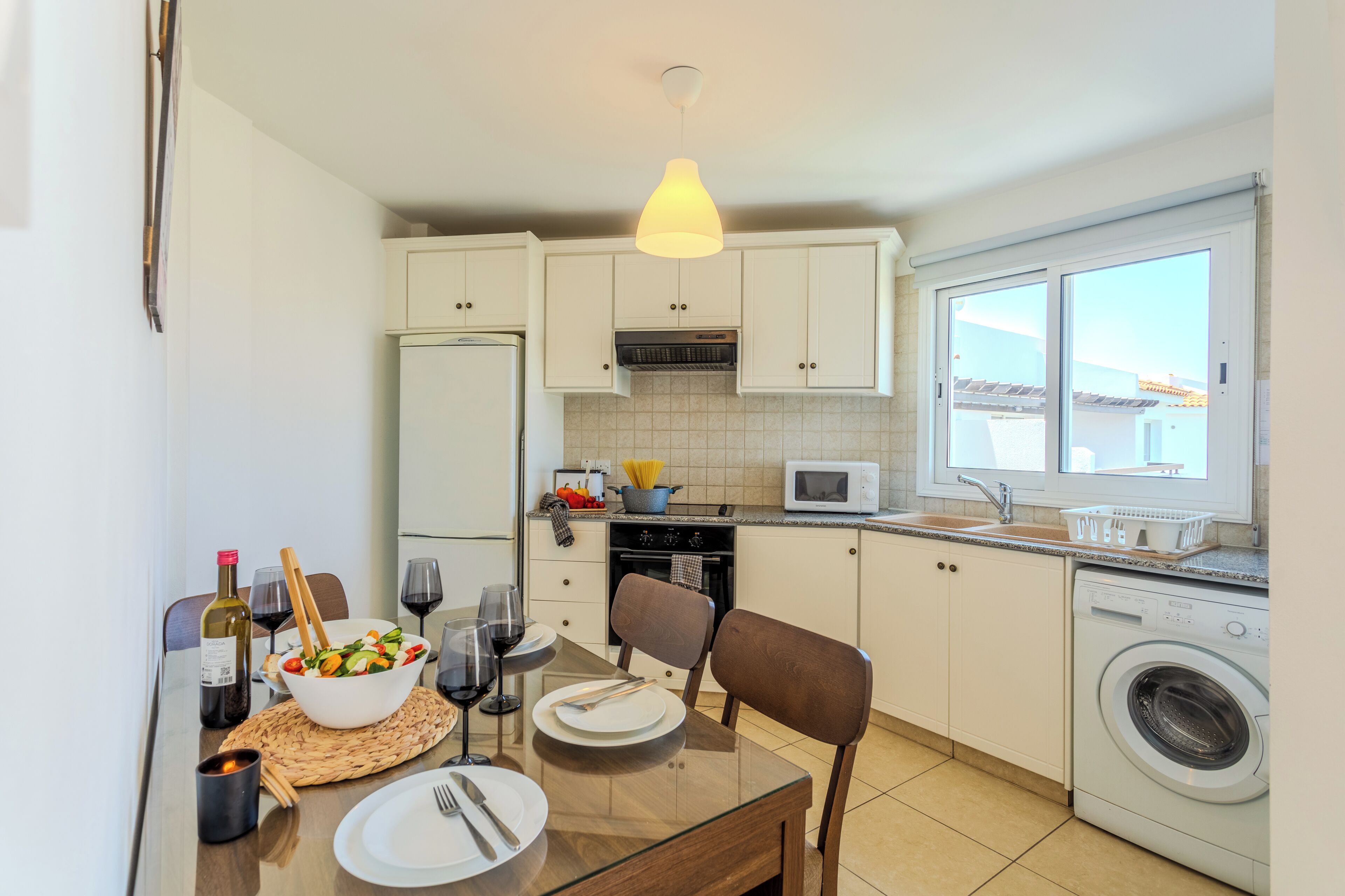 A compact kitchen with white cabinets, a microwave, a washing machine, and a dining area with a table set for two.