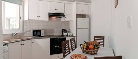 A modern kitchen with white cabinets, a microwave, oven, and a refrigerator. There is a dining table with chairs and a fruit basket.