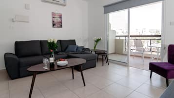 A modern living room with a grey sofa, a small coffee table, and a dining table. There is a balcony with a view of the city.