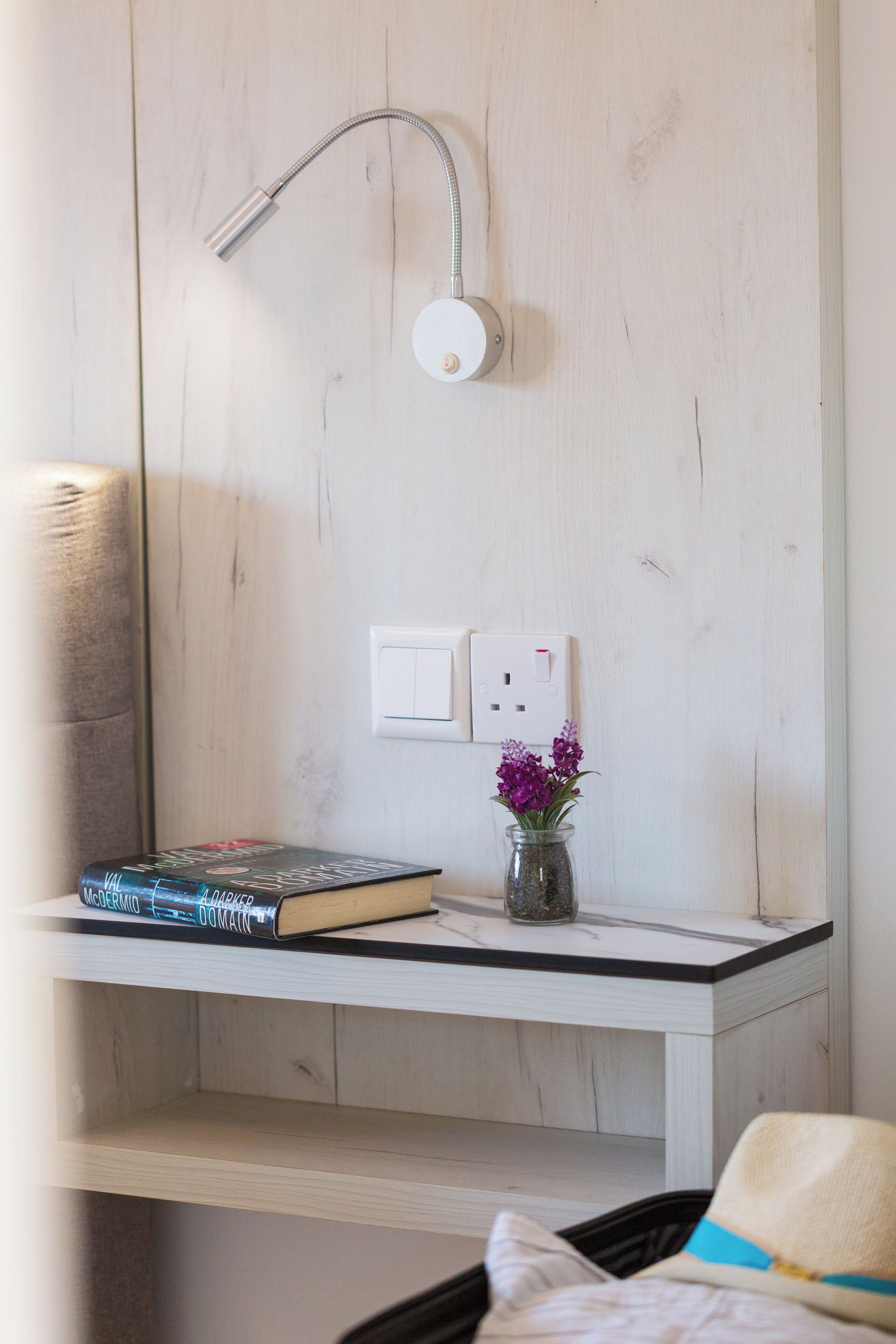Una habitación con pared de madera, un estante blanco con un libro y un jarrón de flores, un cuadro de interruptores blanco y una cama con un sombrero encima.