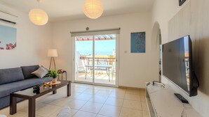 A modern living room with a sofa, coffee table, TV, and a sliding glass door to a patio. - Artemis Cynthia Complex (Paphos)