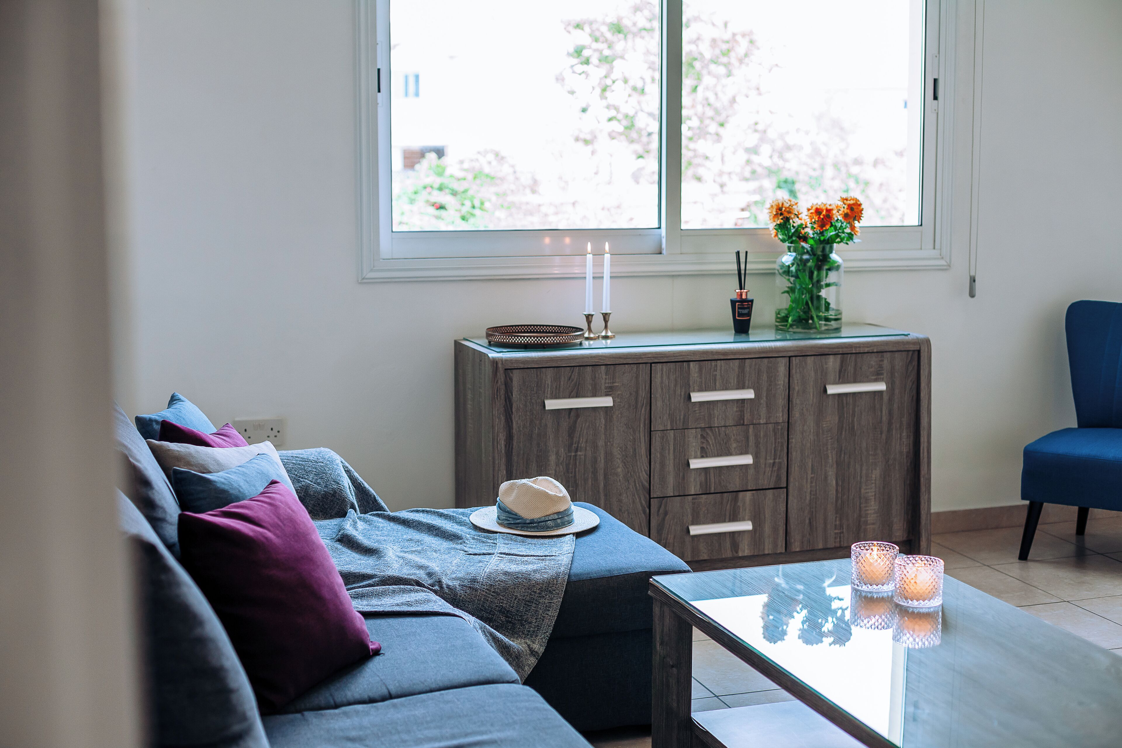 A modern living room with a grey sofa, a blue armchair, and a glass coffee table. There is a wooden cabinet with a vase of flowers and two candles on top.