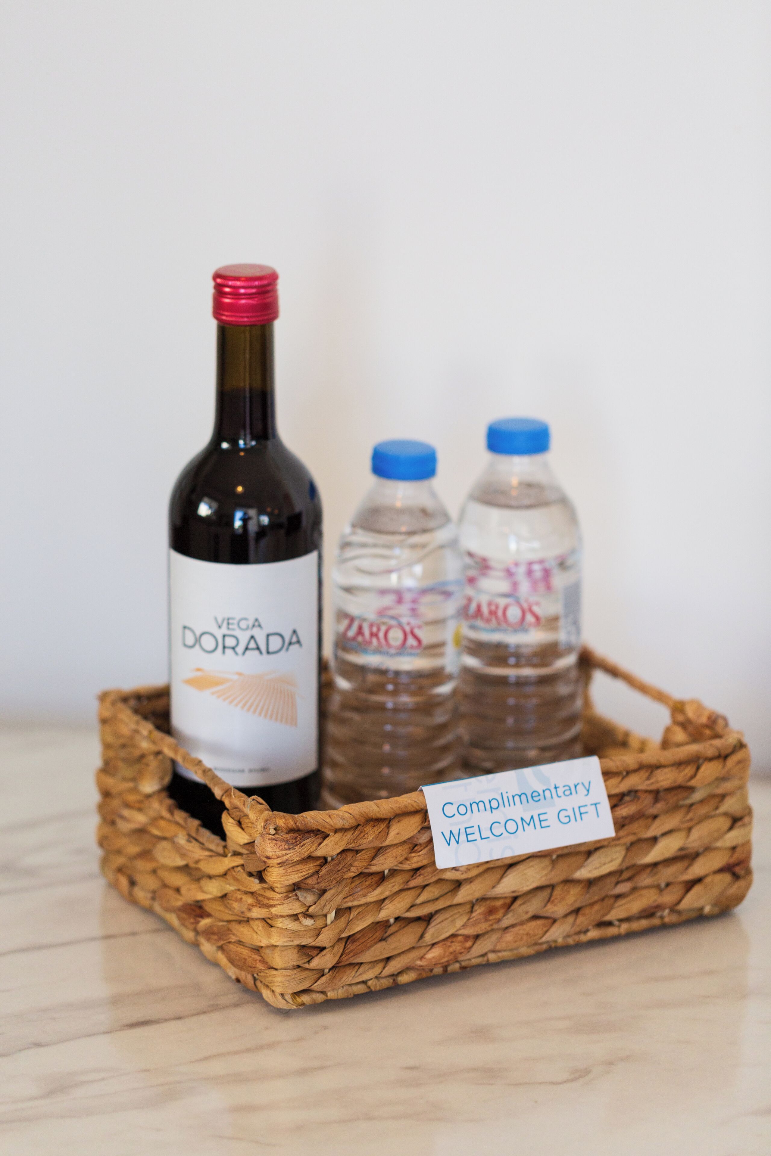 A wicker basket containing a bottle of Vega Dorada wine and two water bottles labeled Zaros. A note on the basket reads "Complimentary Welcome Gift."