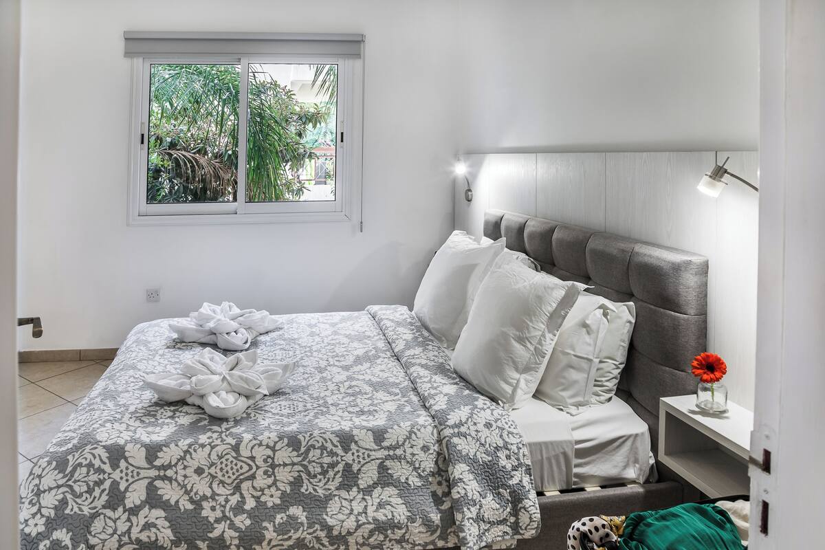 Cama bem arrumada com colcha estampada, mesa de cabeceira com um vaso de flores e janela com vista para o verde.