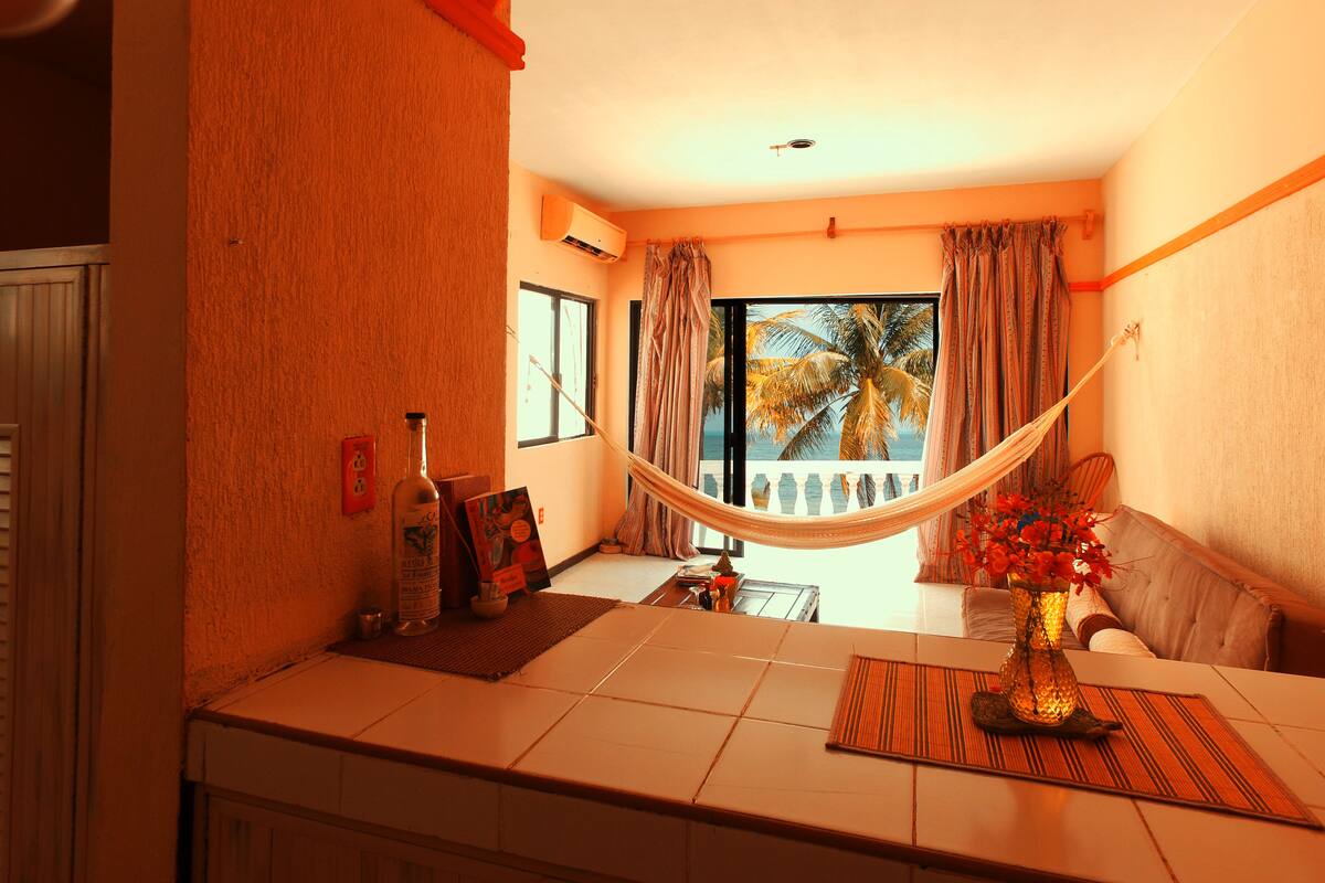 A room with a large window, a hammock, a table with a vase of flowers, and a view of palm trees.