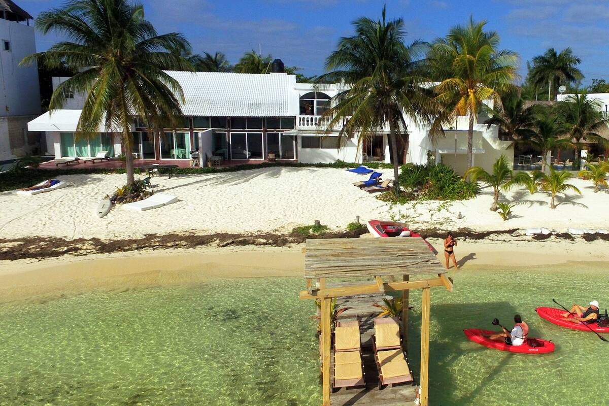 A beach resort with clear water, palm trees, and a wooden walkway leading to a building with large windows.