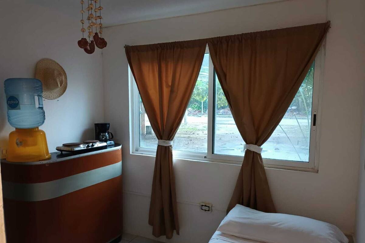 A small room with a bed, a desk with a water dispenser, a coffee maker, and a window with curtains tied back.