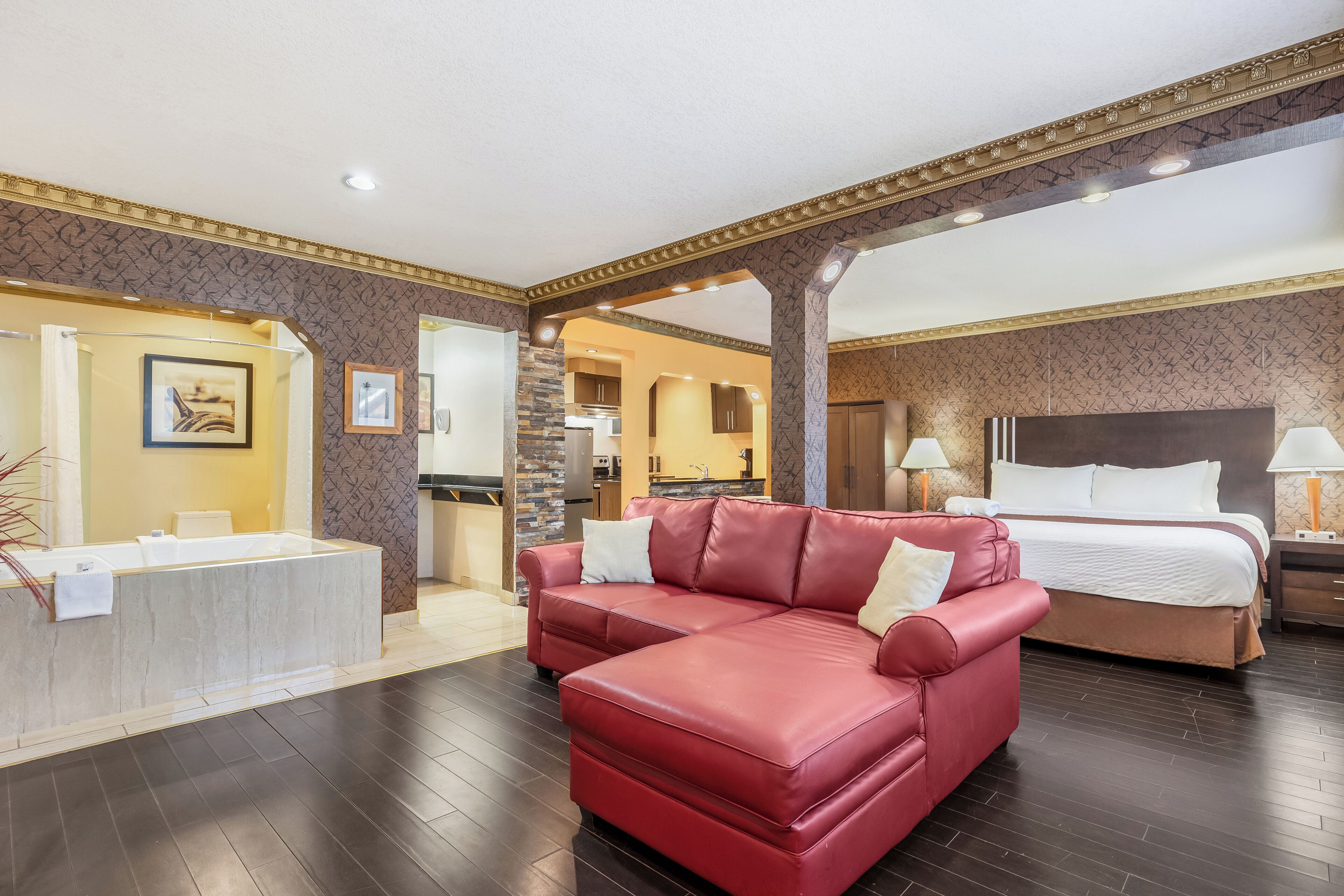 A hotel room with a red leather sofa, a bed with white bedding, a bathroom with a bathtub, and a kitchen area visible through an open doorway.
