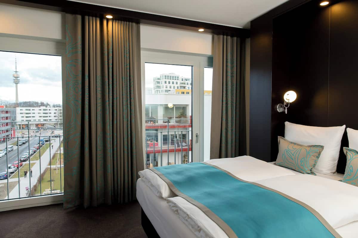 A hotel room with a large bed, a view of a cityscape, and a balcony with a glass railing.