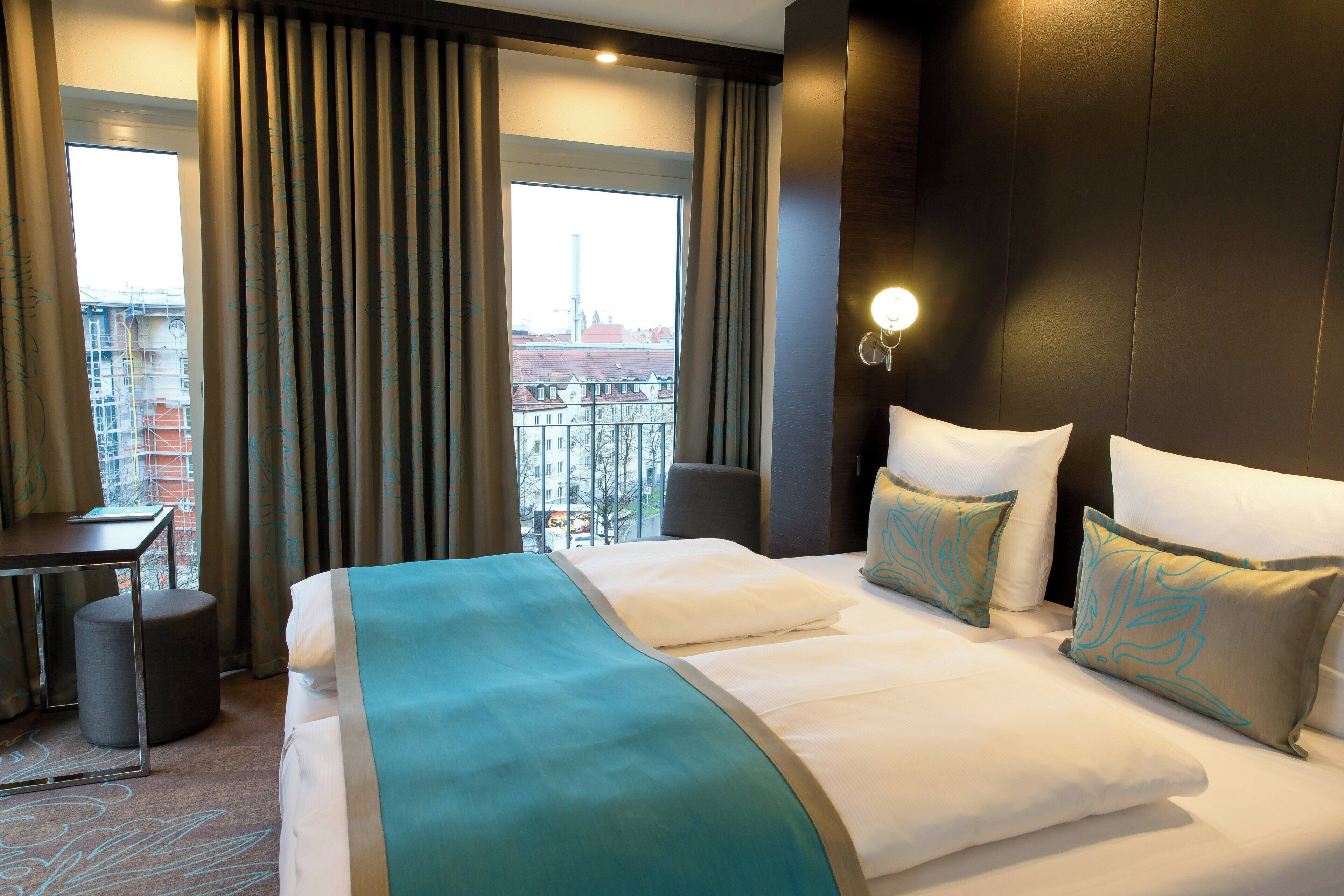 A hotel room with a large bed, a desk, and a view of the cityscape through the window.