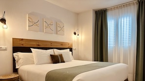 A neatly made bed with a wooden headboard, white bedding, and a green throw blanket. There are two wall-mounted light fixtures and abstract art above the bed. - LUZ Vejer Hospedería (Vejer de la Frontera)