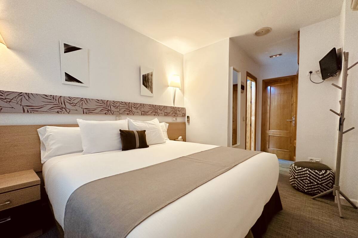 A hotel room with a bed, bedside tables, a lamp, a wall-mounted TV, and a door leading to another room.
