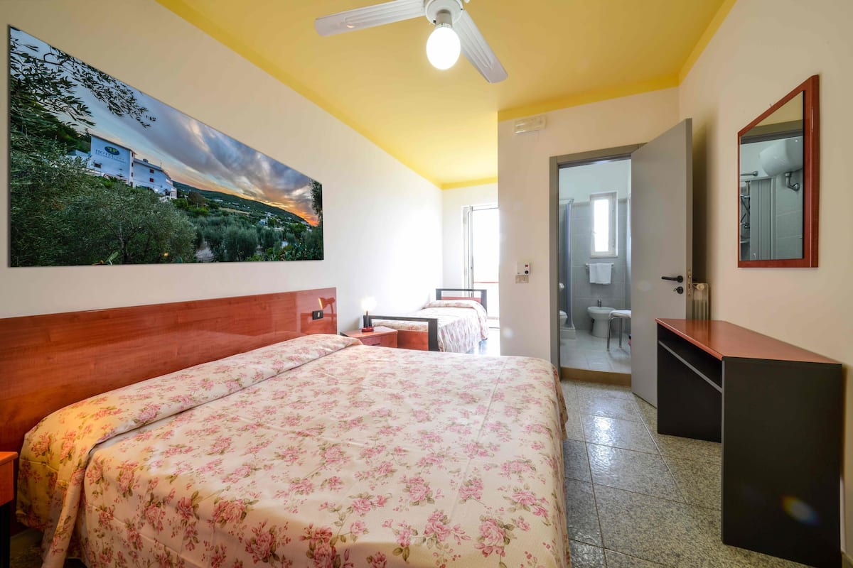 A hotel room with a bed, a painting of a landscape, a ceiling fan, and a bathroom visible through an open door.