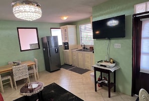 A compact kitchen with a refrigerator, microwave, and a coffee maker. There is a dining table with chairs and a small round table with a decorative item. A television is mounted on the wall. - LeMarida Suites (Speightstown)