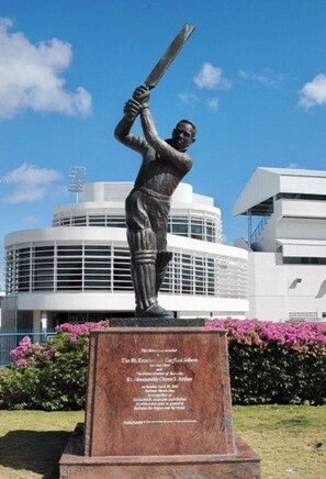 A statue of a cricket player holding a bat, set against a modern building with a circular design. - LeMarida Suites (Speightstown)