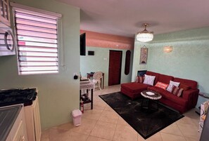 A living room with a red sofa, a small round table, and a black rug. There is a window with blinds, a ceiling fan, and a wall-mounted light fixture. - LeMarida Suites (Speightstown)