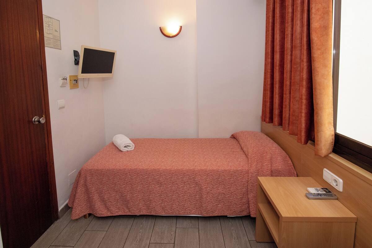 A small, single-bed room with a wooden bedside table, a wall-mounted TV, and a window with curtains.