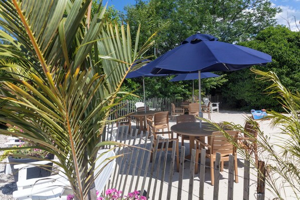 An outdoor patio with tables and chairs, surrounded by a wooden fence and palm trees. - Crosswinds Rehoboth  (Rehoboth Beach)