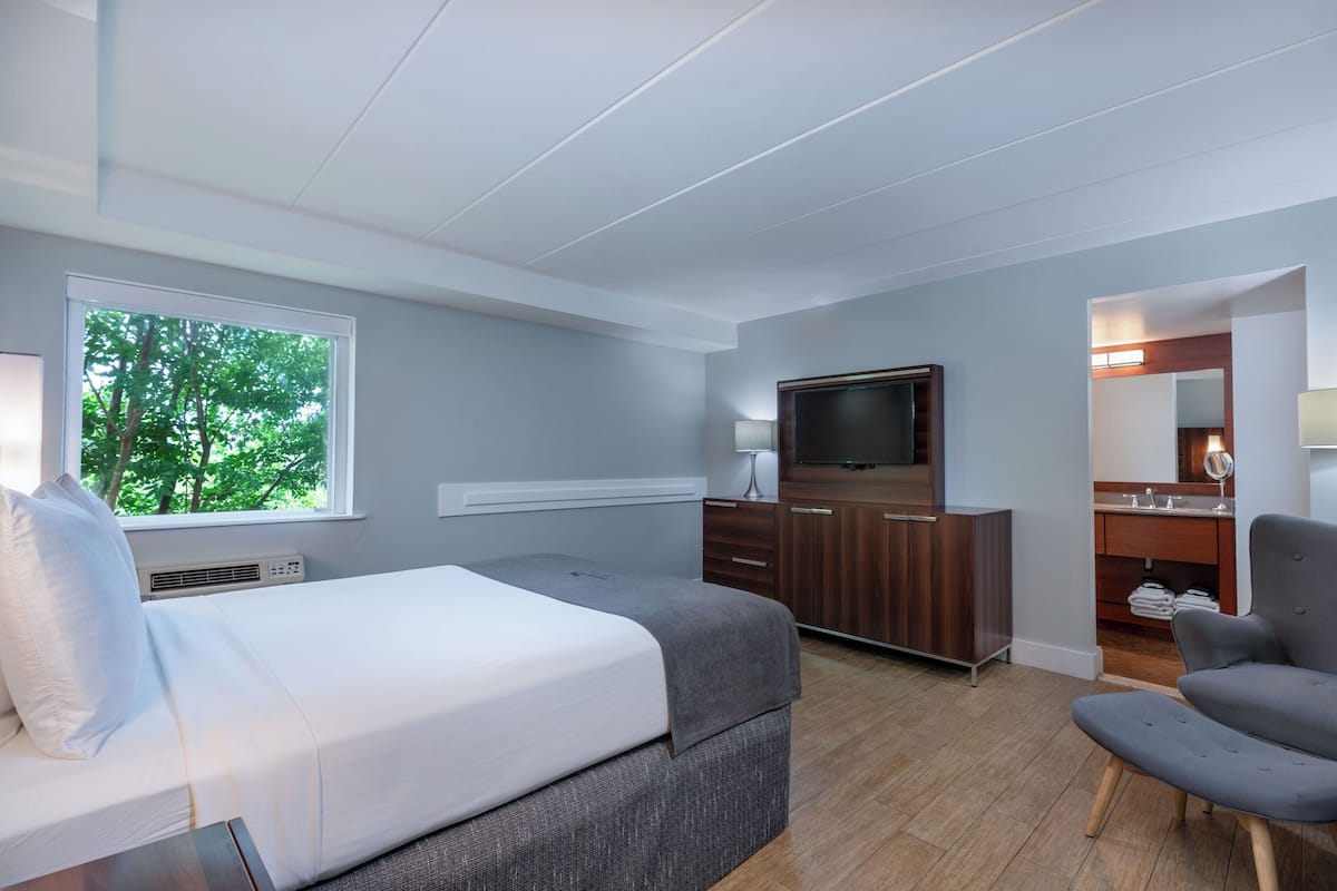 A modern hotel room with a bed, a TV, a desk, a chair, and a bathroom.