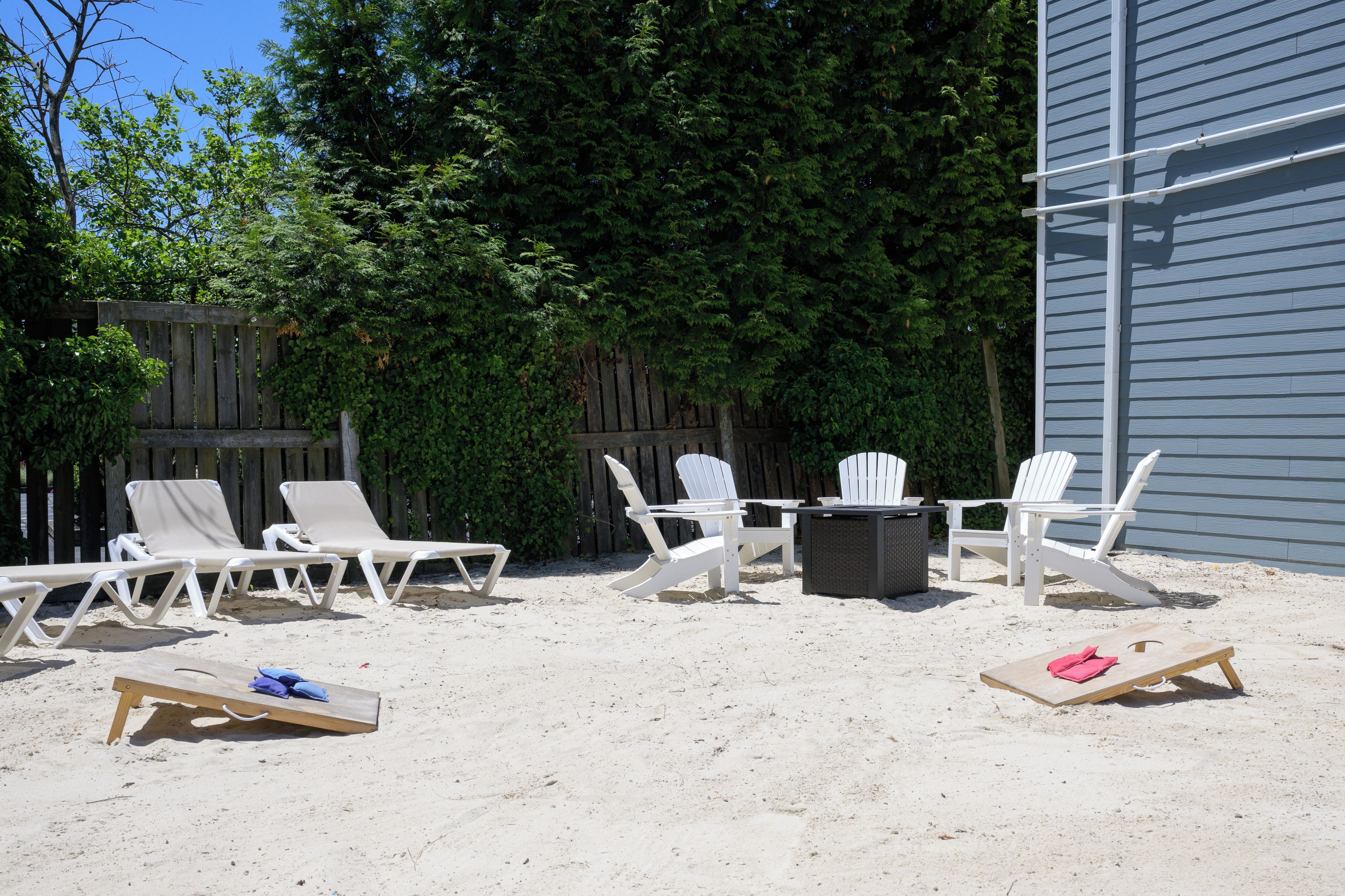 An outdoor seating area with white Adirondack chairs and a table, surrounded by a wooden fence and trees.