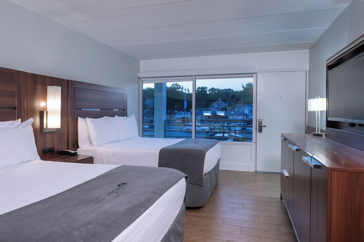 A hotel room with two beds, a TV, a desk, and a large window with a view of a parking lot and buildings.